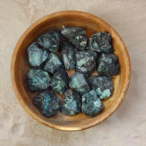 Peut inclure: Un bol en bois rempli de pierres d'émeraude brutes. Les pierres sont vert foncé avec des inclusions vert clair et bleu-vert. Le bol est en bois clair et repose sur une surface texturée de couleur crème.