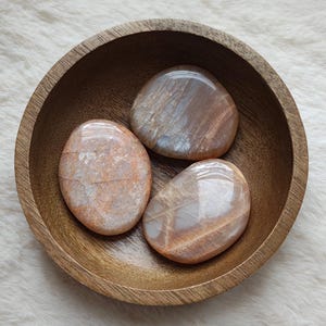 Può includere: Tre pietre di sunstone lisce e ovali a forma di palma in una ciotola di legno. Le pietre sono di colore pesca chiaro con striature bianche e arancioni.