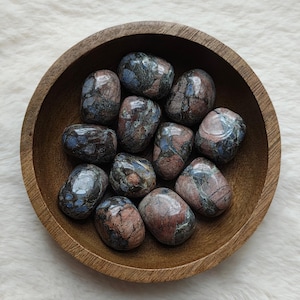 Könnte beinhalten: Eine Holzschale gefüllt mit getrommelten Steinen. Die Steine weisen eine Mischung aus Farben auf, darunter Blau, Braun und Rot. Die Schale besteht aus hellbraunem Holz und liegt auf einer weißen, flauschigen Oberfläche.