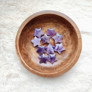May include: A wooden bowl holds several amethyst star-shaped crystals. The crystals vary in shades of purple and are arranged inside the bowl. The bowl is made of wood and has a natural finish. The background is a soft, white, fluffy surface.