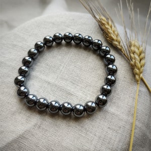 Könnte beinhalten: Ein schwarzes Hämatit-Perlenarmband mit glänzender Oberfläche. Das Armband besteht aus runden Perlen und liegt auf einem hellgrauen Stoffhintergrund.