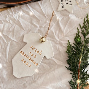Puede incluir: Un adorno de cer&aacute;mica blanca con forma de body de beb&eacute; con letras doradas que dicen "Iyla's 1st Christmas 2023". El adorno tiene una peque&ntilde;a campana dorada y un colgador de cuerda. Tambi&eacute;n se ve un adorno en forma de estrella con "Let it snow".