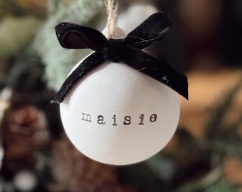 May include: A round white ceramic ornament featuring the name Maisie stamped in black typewriter-style font. The ornament is suspended by a thin twine string and is decorated with a black velvet ribbon tied in a bow at the top.