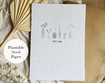 May include: A white card featuring a floral illustration and the text "for you". The card is made from plantable seed paper, which can be planted to grow wildflowers. The card is surrounded by a brown envelope, a white fabric, and a dried palm leaf.
