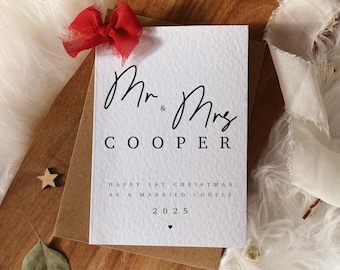 May include: A white card with the text "Mr. & Mrs. COOPER" in black script, with the message "HAPPY 1ST CHRISTMAS AS A MARRIED COUPLE 2025". The card is on a wooden tray with a red bow and gold stars.