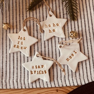 May include: Four white ceramic star ornaments with gold lettering. The ornaments say "let it snow", "ho ho ho", "star bright", and "noel".