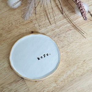 Może przedstawiać: Biała ceramiczna miseczka z pozłacanym rantem i napisem "keys." w czarnych literach. Miseczka leży na jasnym drewnianym tle.