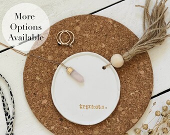 May include: A white ceramic trinket dish with the word "trinkets" in gold lettering. A gold necklace with a pink crystal pendant and two gold rings rest on the dish. The dish sits on a round cork coaster. The text "More Options Available" is in the upper left.