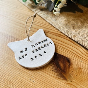 May include: A white ceramic cat-shaped ornament with the text "my humans are engaged 2024" stamped in black. The ornament hangs by a twine loop. The background includes a burlap cloth and a floral arrangement.