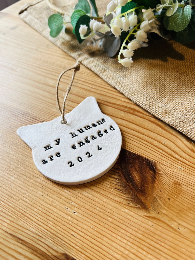 May include: A white ceramic cat-shaped ornament with the text "my humans are engaged 2024" stamped in black. The ornament hangs by a twine loop. The background includes a burlap cloth and a floral arrangement.