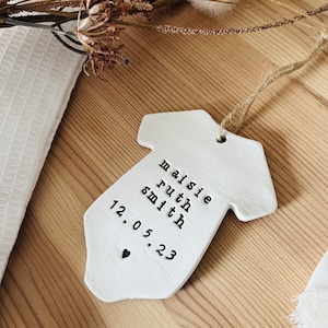 May include: White ceramic baby onesie ornament with the text "Maisie Ruth Smith 12.05.23" and a small black heart.