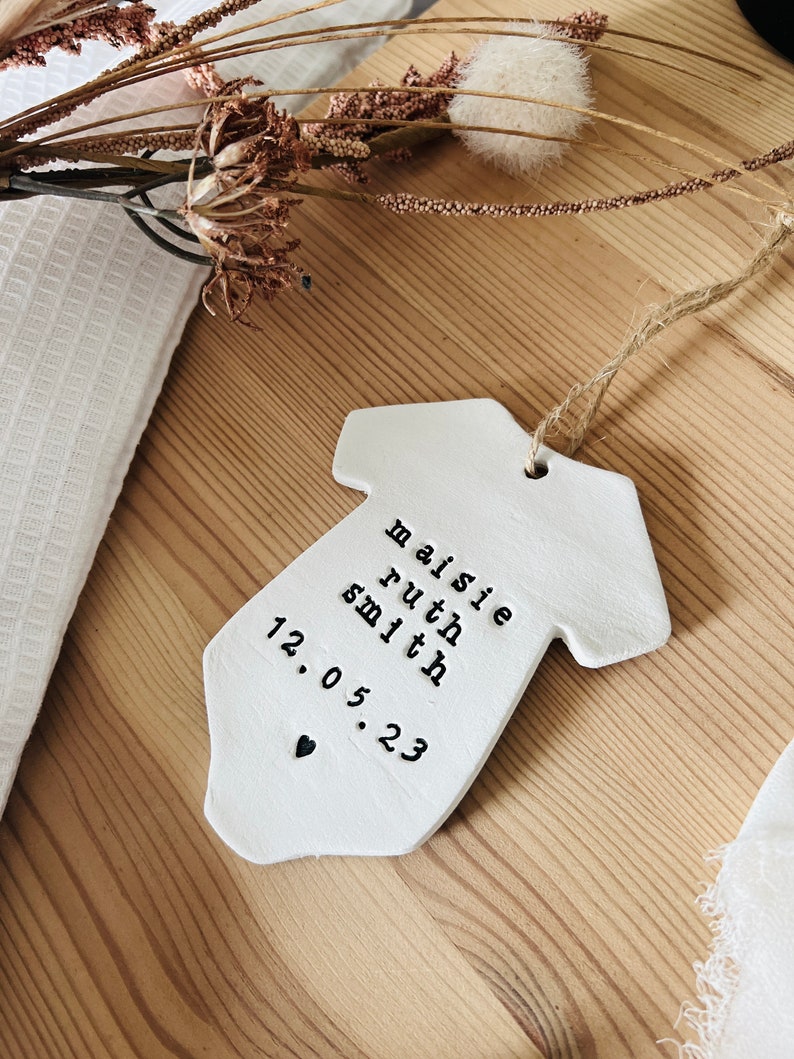 May include: White ceramic baby onesie ornament with the text "Maisie Ruth Smith 12.05.23" and a small black heart.
