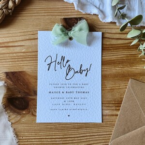May include: A light blue baby shower invitation with a green ribbon bow and the text "Hello Baby!"  Please join us for a baby shower celebrating Maisie & Baby Thomas. Saturday 18th May 2023 @ 12PM. Cafe Nine, Hauley. RSVP Claire 07967452312.