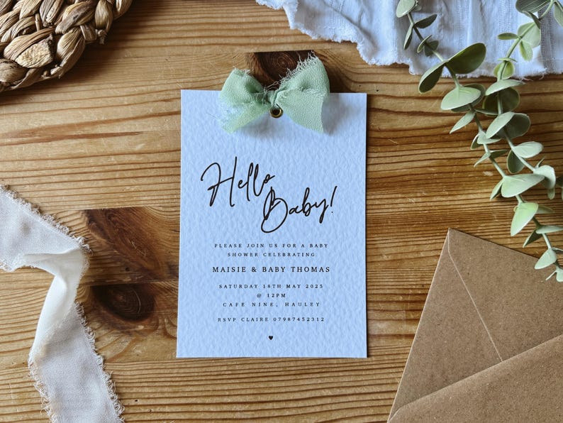 May include: A light blue baby shower invitation with a green ribbon bow and the text "Hello Baby!"  Please join us for a baby shower celebrating Maisie & Baby Thomas. Saturday 18th May 2023 @ 12PM. Cafe Nine, Hauley. RSVP Claire 07967452312.