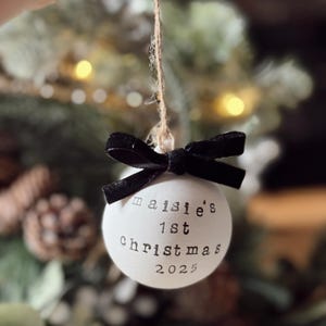 May include: A white Christmas ornament with the text "maisie's 1st Christmas 2025" in black. The ornament is tied with a black velvet bow and suspended by a twine string. The background is blurred with Christmas tree branches.