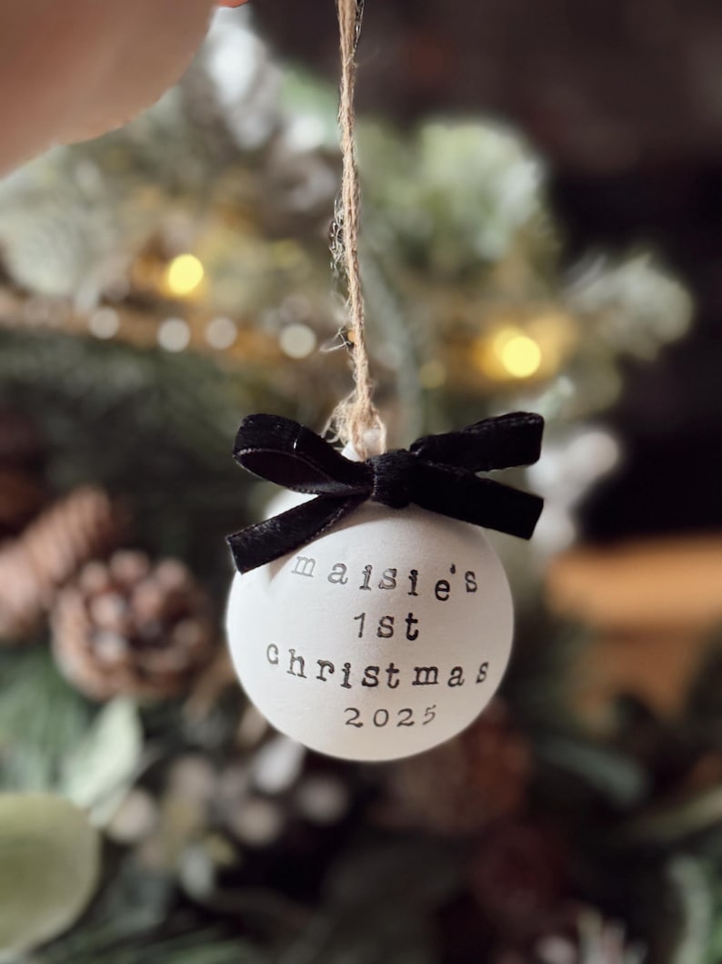 May include: A white Christmas ornament with the text "maisie's 1st Christmas 2025" in black. The ornament is tied with a black velvet bow and suspended by a twine string. The background is blurred with Christmas tree branches.