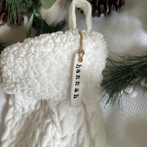 May include: White knitted stocking with a fluffy white cuff and a white tag that says "hannah" hanging from it.