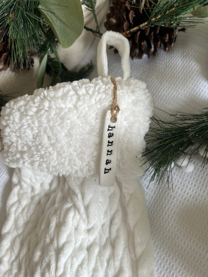 May include: White knitted stocking with a fluffy white cuff and a white tag that says "hannah" hanging from it.