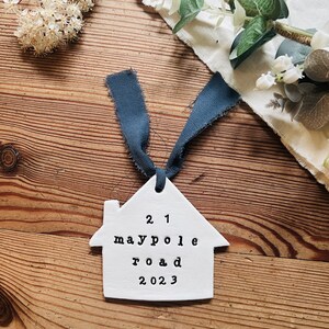 May include: White ceramic house ornament with a blue ribbon. The ornament has the text "21 maypole road 2023" on it.