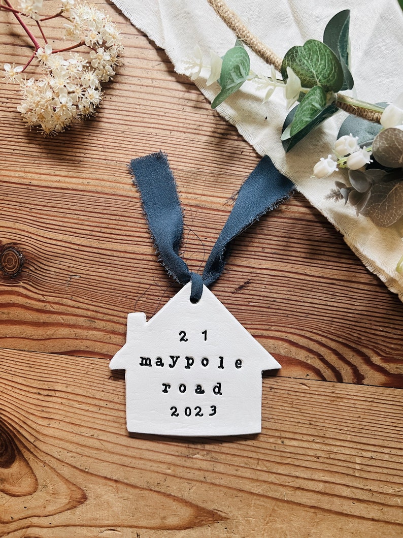 May include: White ceramic house ornament with a blue ribbon. The ornament has the text "21 maypole road 2023" on it.