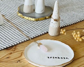 May include: A collection of handmade jewelry holders and a trinket dish. The holders are cone-shaped, painted white with a beige base. A round white dish with gold trim has the word "trinkets" printed on it. A necklace and rings are also present.