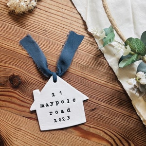 May include: White ceramic house ornament with the text "21 Maypole Road 2023" and a blue ribbon.