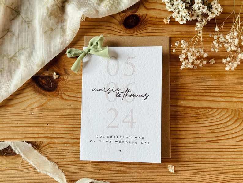 May include: A white card with the text "05 06 24" and "Maisie & Thomas" in black script. The card also says "Congratulations on your wedding day" in black text. The card is on a wooden surface with a green ribbon tied on top.