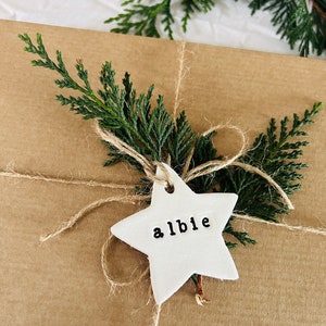 May include: A wrapped gift in brown paper, tied with twine and decorated with evergreen sprigs. A white star-shaped tag with the name "albie" is attached. The background includes a pine cone and white paper.