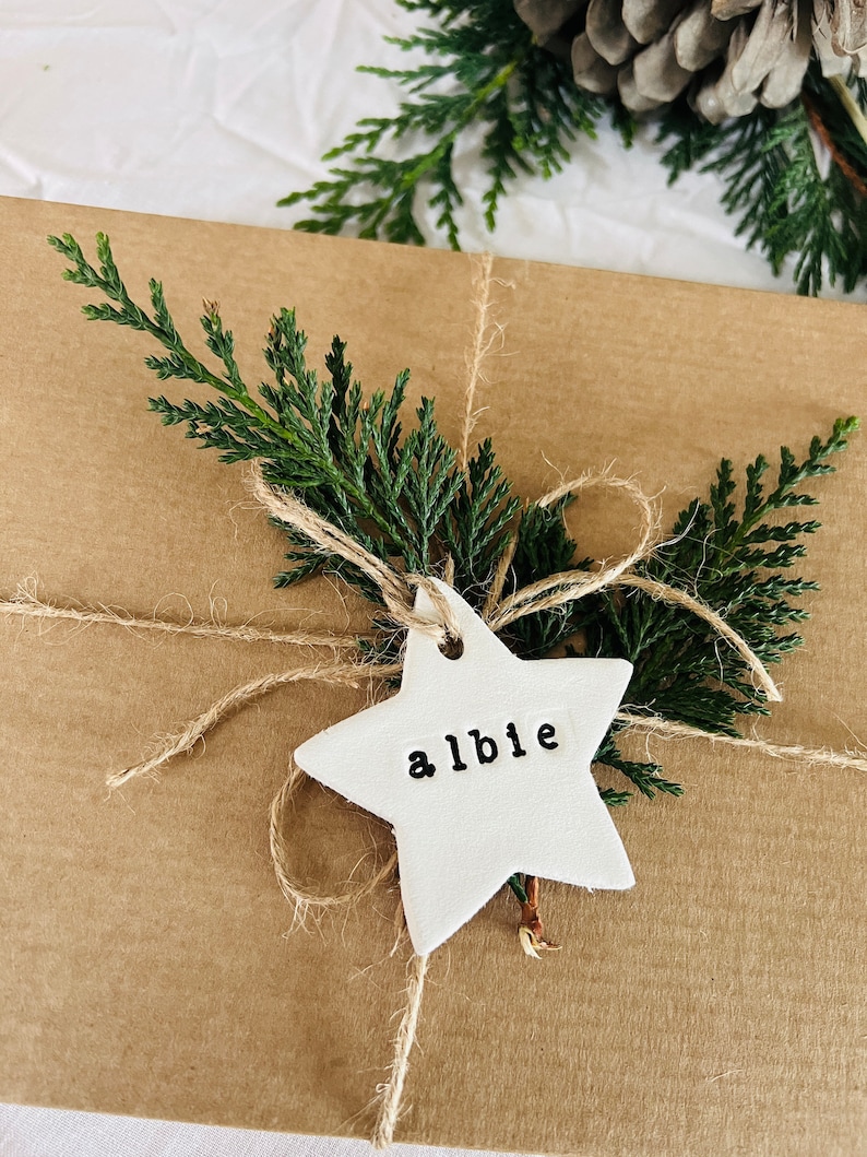 May include: A wrapped gift in brown paper, tied with twine and decorated with evergreen sprigs. A white star-shaped tag with the name "albie" is attached. The background includes a pine cone and white paper.