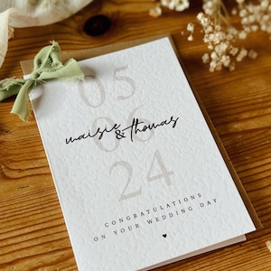 May include: A white card with brown text and a green ribbon. The text reads "05 06 Maisie & Thomas 24 Congratulations on your wedding day".