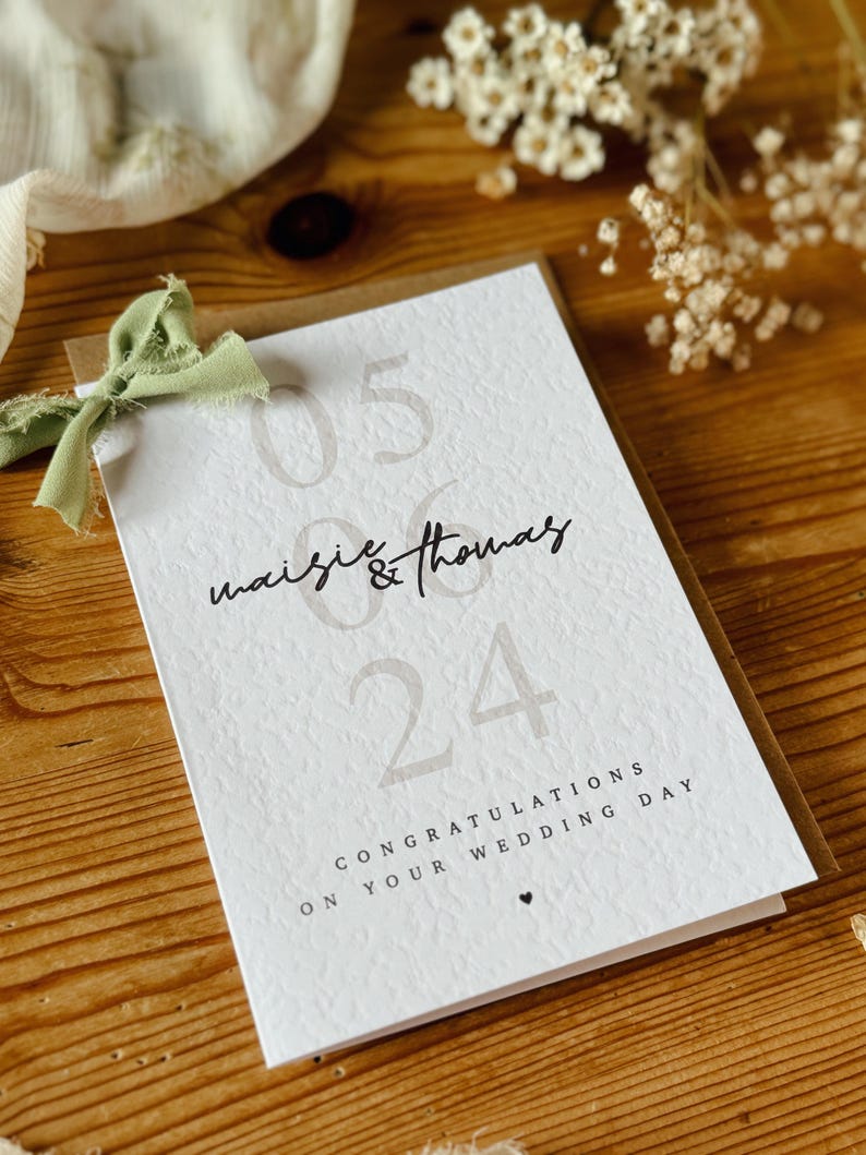 May include: A white card with brown text and a green ribbon. The text reads "05 06 Maisie & Thomas 24 Congratulations on your wedding day".
