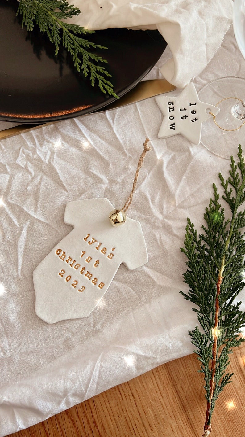 Puede incluir: Un adorno de cer&aacute;mica blanca con forma de body de beb&eacute; con letras doradas que dicen "Iyla's 1st Christmas 2023". El adorno tiene una peque&ntilde;a campana dorada y un colgador de cuerda. Tambi&eacute;n se ve un adorno en forma de estrella con "Let it snow".