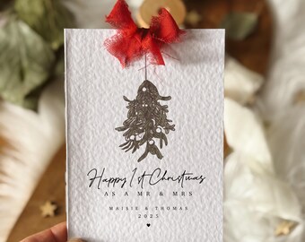 May include: A white card with the text "Happy 1st Christmas as a Mr & Mrs" and names. A brown mistletoe design hangs below the text. A red bow is attached to the top of the card. The card is held by a person's hand.