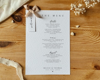 May include: A white wedding menu card with black text. The menu includes starter, main, and dessert options. The card is decorated with a small, light brown ribbon bow. The text at the bottom of the card reads "Maisie & Thomas 5th June 2025".