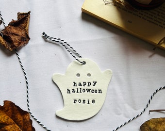May include: A white ceramic ghost-shaped ornament with the words "happy halloween rosie" stamped on it. The ornament is strung with black and white twine and is surrounded by dried leaves and a book.