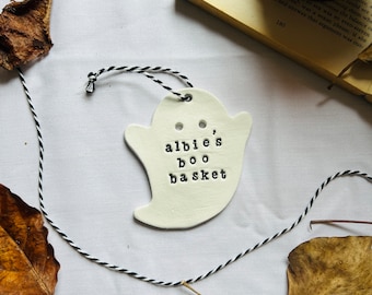 May include: A white ceramic ghost-shaped ornament with the text "albie's boo basket" stamped on it. The ornament is strung with black and white twine. The image includes dried leaves and a book.