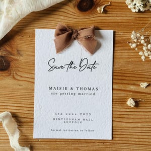 May include: Save the Date card with a brown bow on top. The card is white with black text that reads "Save the Date", "Maisie & Thomas", "are getting married", "5th June 2025", "Hintlesham Hall", "Suffolk", and "formal invitation to follow".