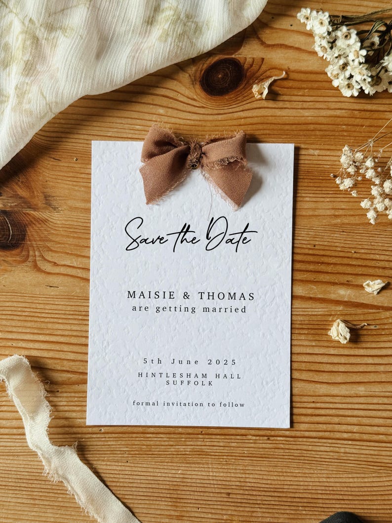 May include: Save the Date card with a brown bow on top. The card is white with black text that reads "Save the Date", "Maisie & Thomas", "are getting married", "5th June 2025", "Hintlesham Hall", "Suffolk", and "formal invitation to follow".