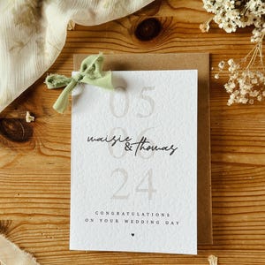 May include: A white card with the text "05 Maisie & Thomas 24" and the message "Congratulations on your wedding day". The card is decorated with a green ribbon.