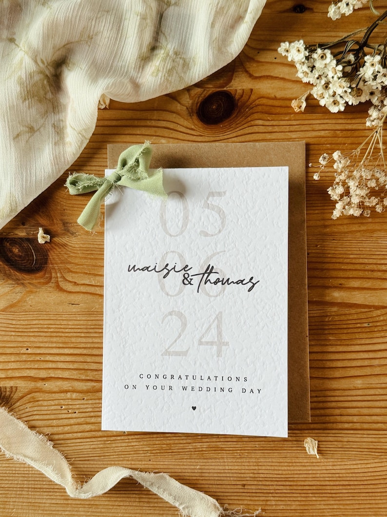 May include: A white card with the text "05 Maisie & Thomas 24" and the message "Congratulations on your wedding day". The card is decorated with a green ribbon.