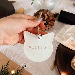 May include: A white cat-shaped ornament with the name "millie" stamped in silver letters. The ornament hangs from a twine string with a silver bell. The ornament is held over a table setting with a brown box and greenery.
