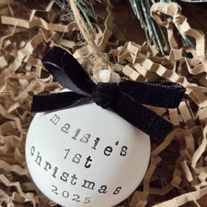 May include: White Christmas ornament with the text "maisie's 1st christmas 2025" in black lettering. The ornament has a black velvet bow and is suspended by a twine string. The background is brown paper and evergreen branches.