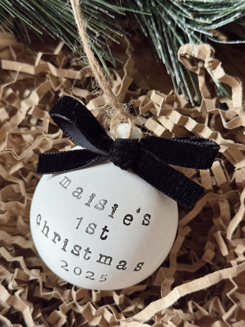 May include: White Christmas ornament with the text "maisie's 1st christmas 2025" in black lettering. The ornament has a black velvet bow and is suspended by a twine string. The background is brown paper and evergreen branches.