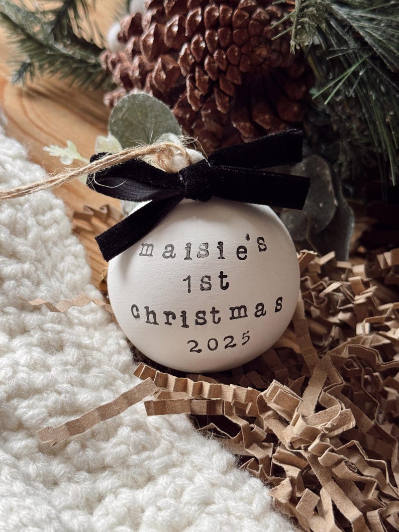 May include: A white Christmas ornament with the text "maisie's 1st christmas 2025" printed in black. The ornament is tied with a black velvet bow and twine. The background includes pine needles and a pine cone.