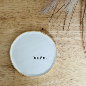 May include: White ceramic dish with a gold rim, featuring the word "keys." on a light wood surface.