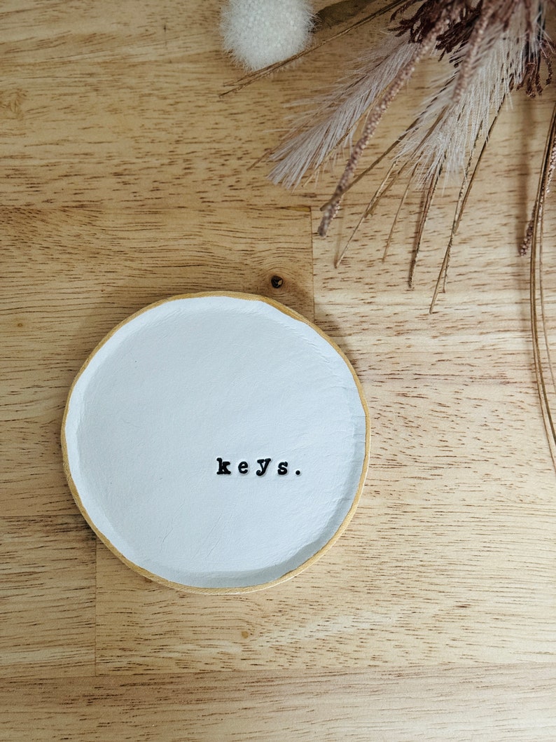 May include: White ceramic dish with a gold rim, featuring the word "keys." on a light wood surface.