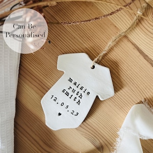 May include: White ceramic baby onesie ornament with a string for hanging. The onesie has the text "maisie ruth smith 12.05.23" written on it.