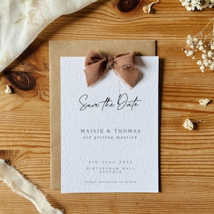 May include: Save the Date card with a brown ribbon bow, featuring the text "Save the Date", "Maisie & Thomas are getting married", "5th June 2025", "Histlesham Hall Suffolk", and "formal invitation to follow".