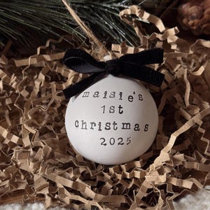 May include: A white Christmas ornament with the text "maisie's 1st Christmas 2025" in black lettering. The ornament is adorned with a black velvet bow and a twine string for hanging. The ornament is nestled in brown paper and greenery.