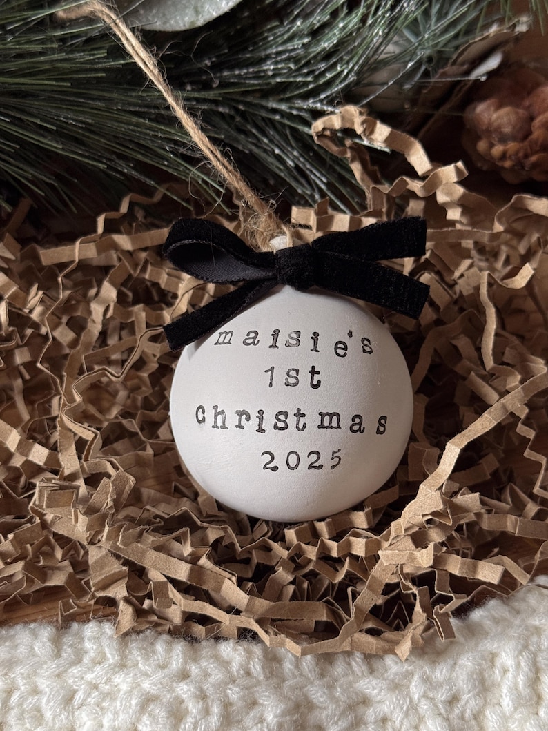 May include: A white Christmas ornament with the text "maisie's 1st Christmas 2025" in black lettering. The ornament is adorned with a black velvet bow and a twine string for hanging. The ornament is nestled in brown paper and greenery.
